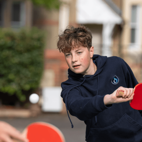 Person playing table tennis outdoors, wearing a dark hoodie with a small droplet logo on the chest. The individual is holding a red paddle and appears to be in mid-action, hitting a white ping pong ball. Another red paddle is partially visible in the foreground. The background shows a blurred building and greenery.